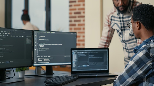 Free Photo focused software developer writing code on laptop looking at multiple screens with programming language is interrupted by colleague coder asking for advice. programmers doing online cloud computing.