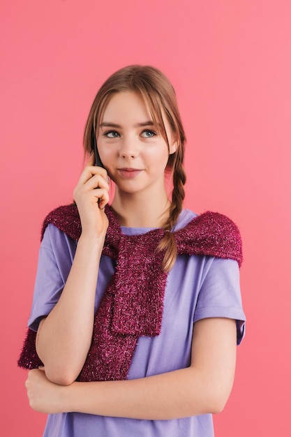 Free Photo young beautiful girl with two braids in lilac t shirt and sweater on shoulders talking on cellphone while happily looking aside over pink background