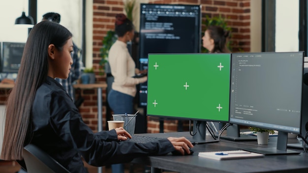Photo asian programer writing code in front of computer with green screen chroma key mockup in software development agency. coder computing big data with team colleagues working online cloud computing.