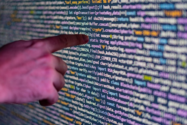 Photo closeup of a pointing hand over a large glowing display screen filled with colorful code possibly representing computer programming or data visualization