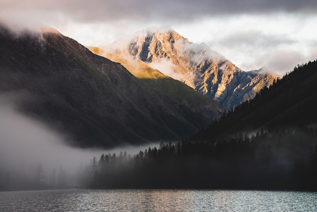 Photo high gold mountain peak and many low clouds above mountain lake at sunrise. dense fog above water and forest in golden hour. atmospheric highland landscape at early morning. alpine relaxing scenery.