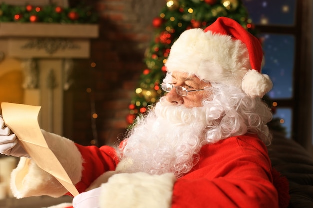 Photo portrait of happy santa claus sitting at his room at home near christmas tree and reading christmas letter or wish list.