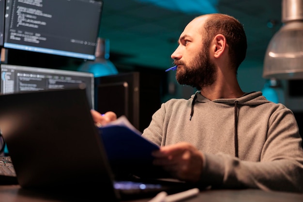 Photo web developer analyzing source code with papers and computer, taking notes and working on web development. male programmer comparing files with online server data after hours.